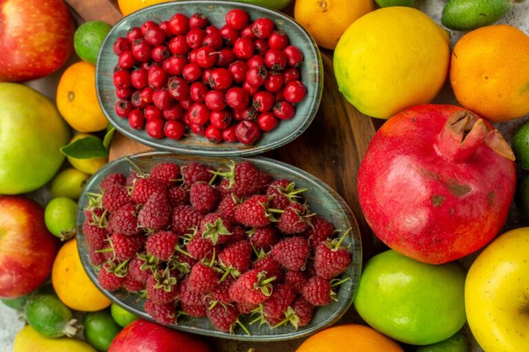 A variety of fresh fruits including pomegranate, raspberries, cranberries, apples, and citrus fruits are arranged on a table.