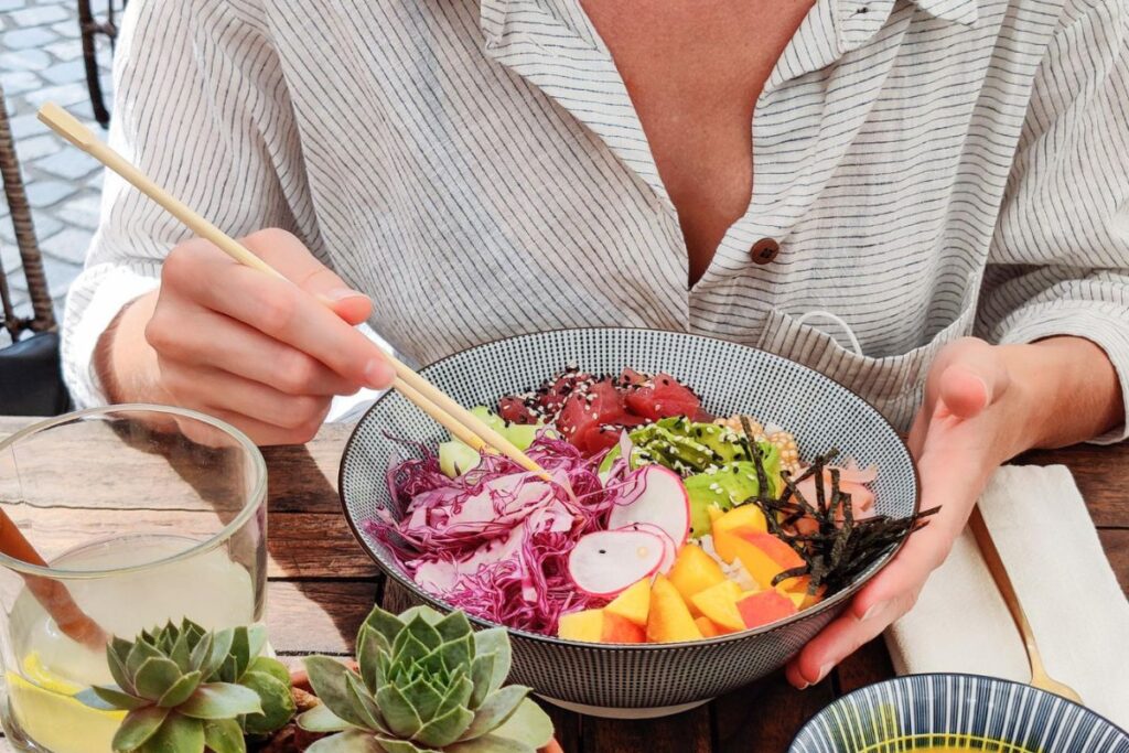 A person wearing a striped shirt is holding a bowl of colorful salad with purple cabbage, mango, avocado, radish, and other ingredients, using chopsticks. A glass of drink and succulents are nearby. This vibrant dish is perfect for those exploring candida diet recipes.