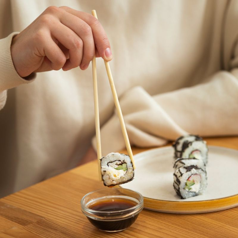 A person holding a piece of sushi with chopsticks dips it into a small bowl of soy sauce. The plate with more sushi pieces is on a wooden table.