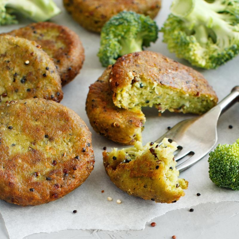 Four cooked vegetable patties on parchment paper with a fork breaking one open, surrounded by broccoli florets.