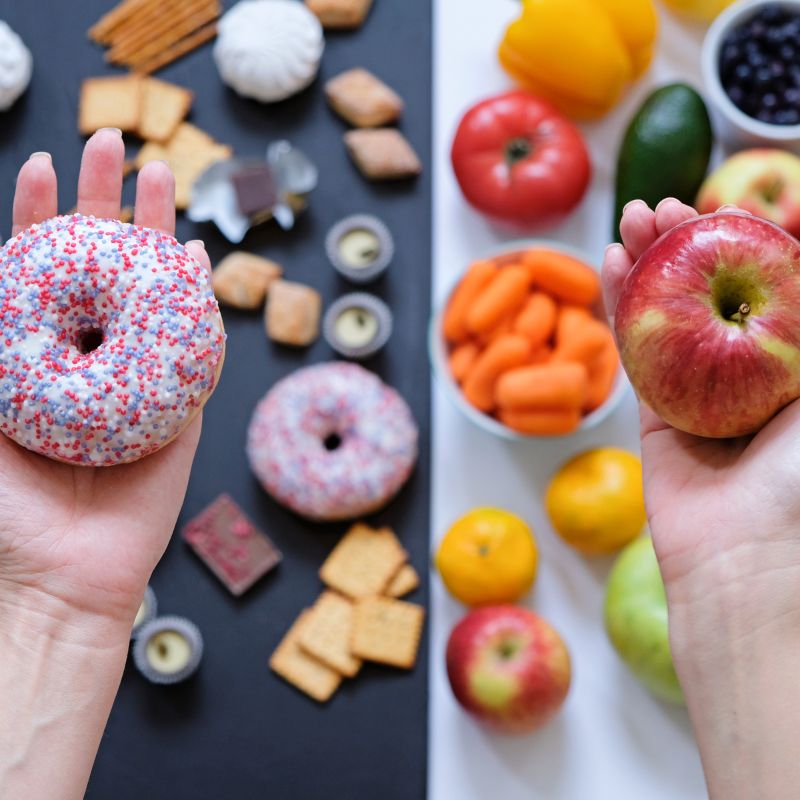 A person holds a donut in one hand and an apple in the other. Various snacks including fruits, vegetables, and sweets are spread out on a black and white surface in the background.