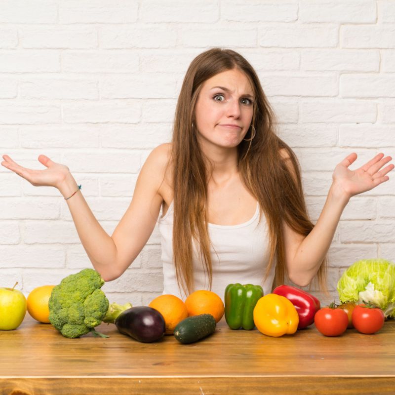 A person with long hair in a white tank top shrugs with arms raised, standing behind a wooden table filled with various fruits and vegetables.