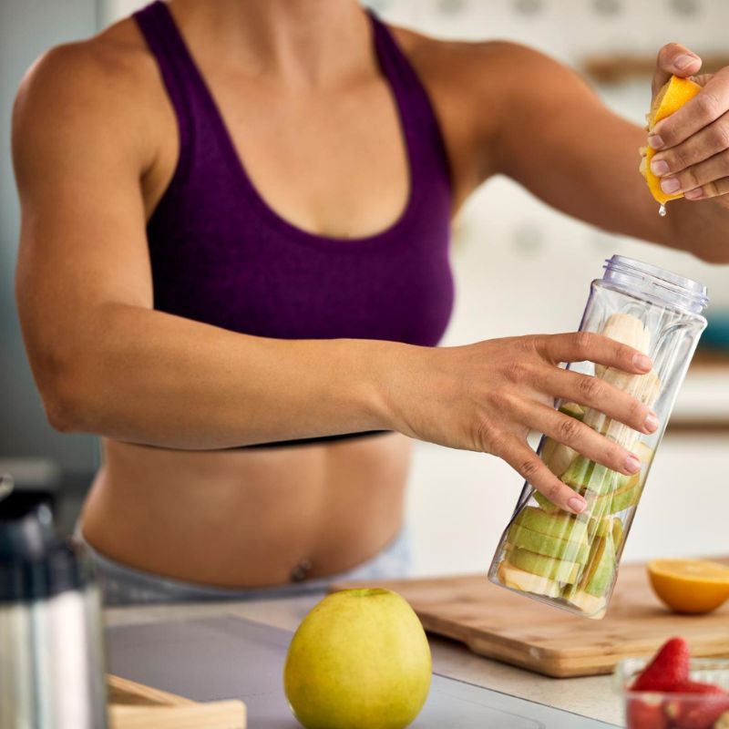 A person in a purple sports top squeezes a lemon into a bottle filled with apple slices in a kitchen setting, crafting one of their favorite healthy drinks.