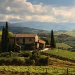 A stone farmhouse with a red-tiled roof stands amidst a lush vineyard on a hillside. Tall cypress trees are nearby, and mountains rise in the distance under a partly cloudy sky.