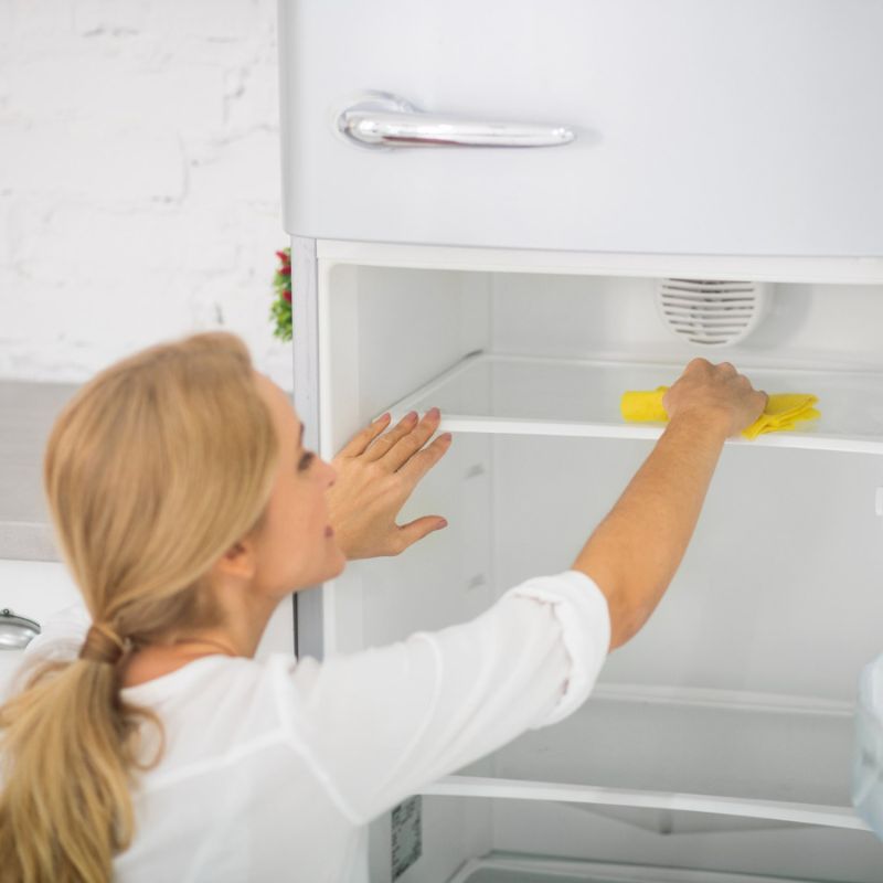 A person is cleaning the interior of an empty refrigerator with a yellow cloth in a kitchen.