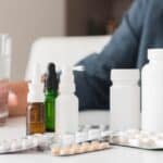 Person taking medication with a glass of water surrounded by various medicines and supplements.