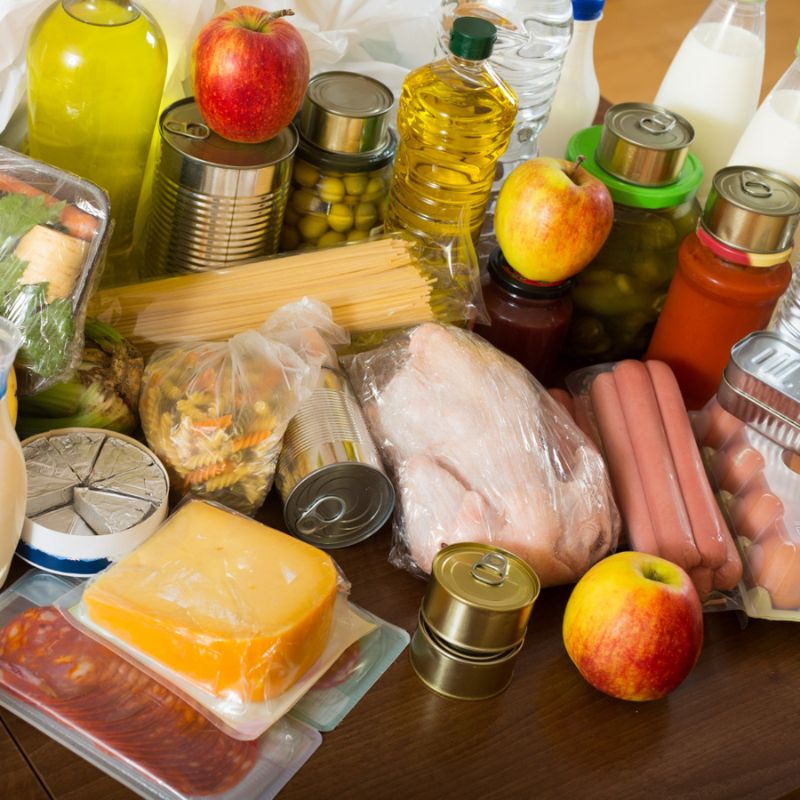 A variety of groceries, including fresh fruits, vegetables, pasta, meat, canned goods, bottles of oil and milk, cheese, and delicatessen meats, are displayed on a wooden table.