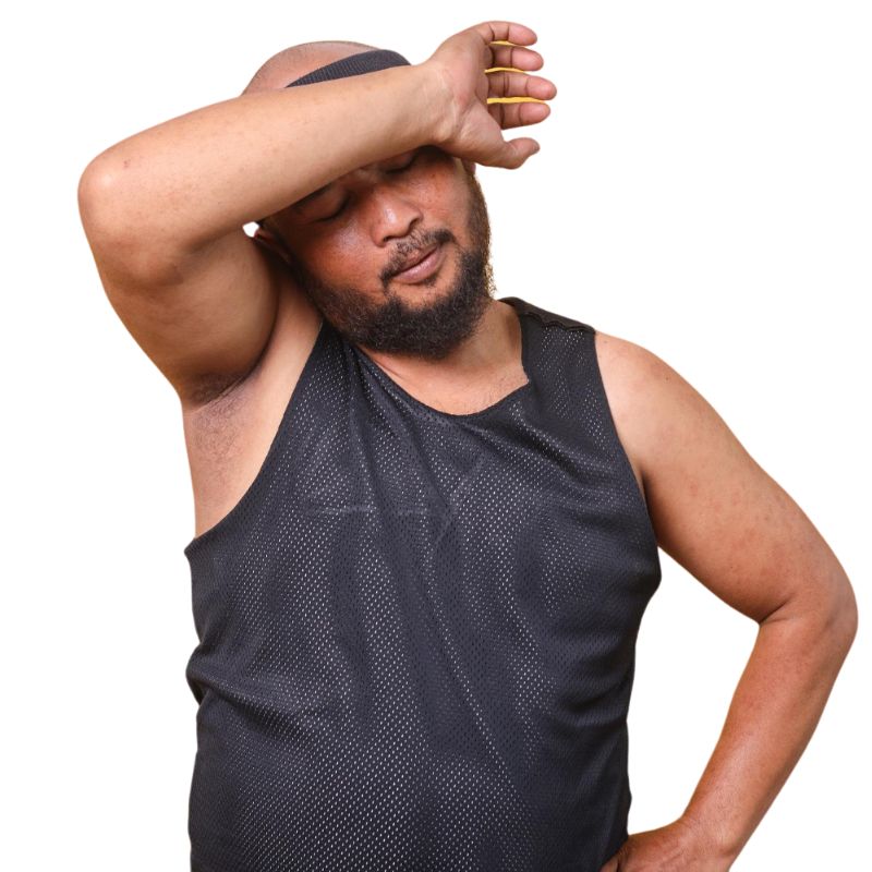 A man in a black sleeveless shirt wipes sweat from his forehead with his arm, wondering if he's experiencing the discomfort of jock itch.