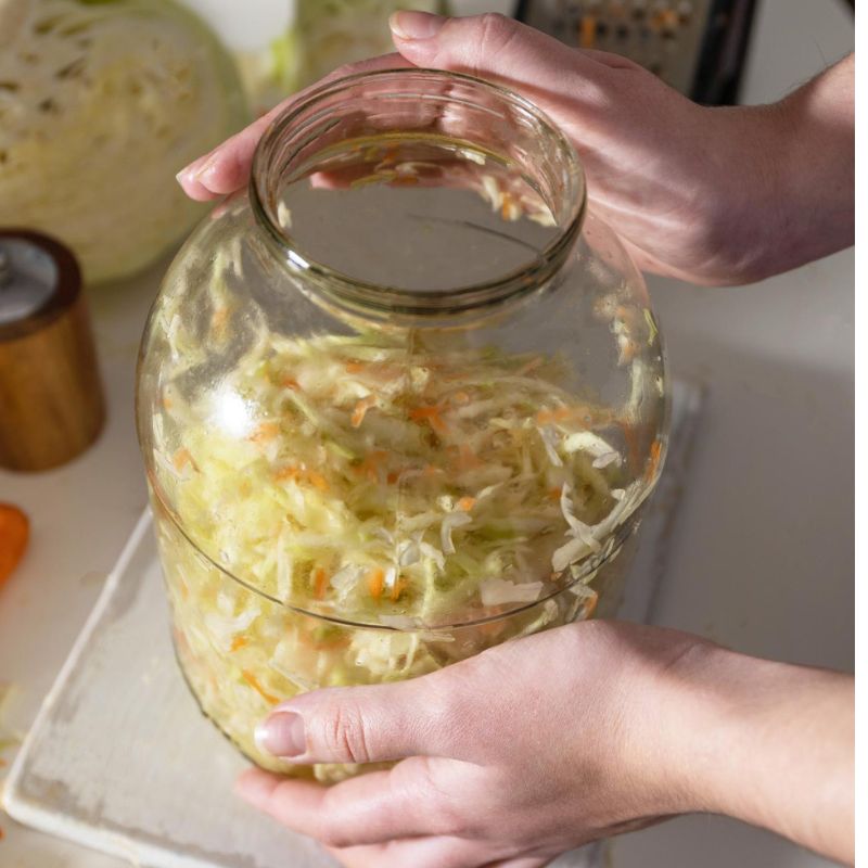 Hands holding a glass jar filled with shredded cabbage and carrots, likely prepared for fermentation. A cutting board and kitchen utensils are visible in the background.