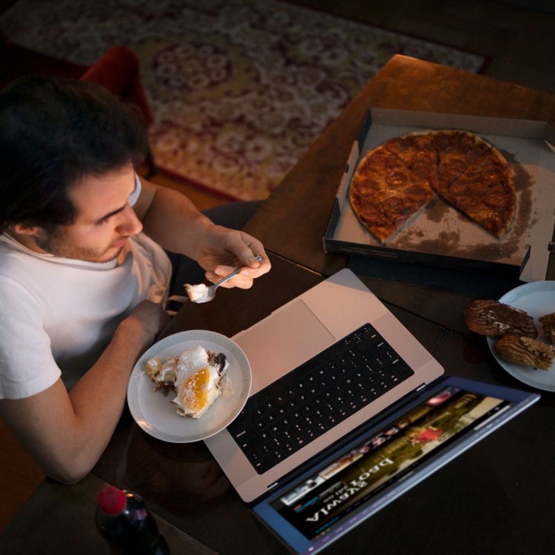 Man eating dessert while working on a laptop, with a pizza box and a drink on a coffee table in a dimly lit room.