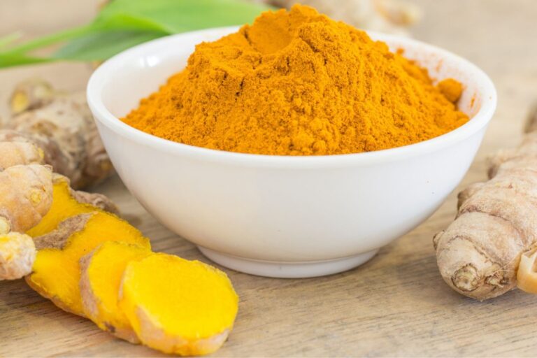A bowl of turmeric powder with fresh turmeric roots alongside on a wooden surface.