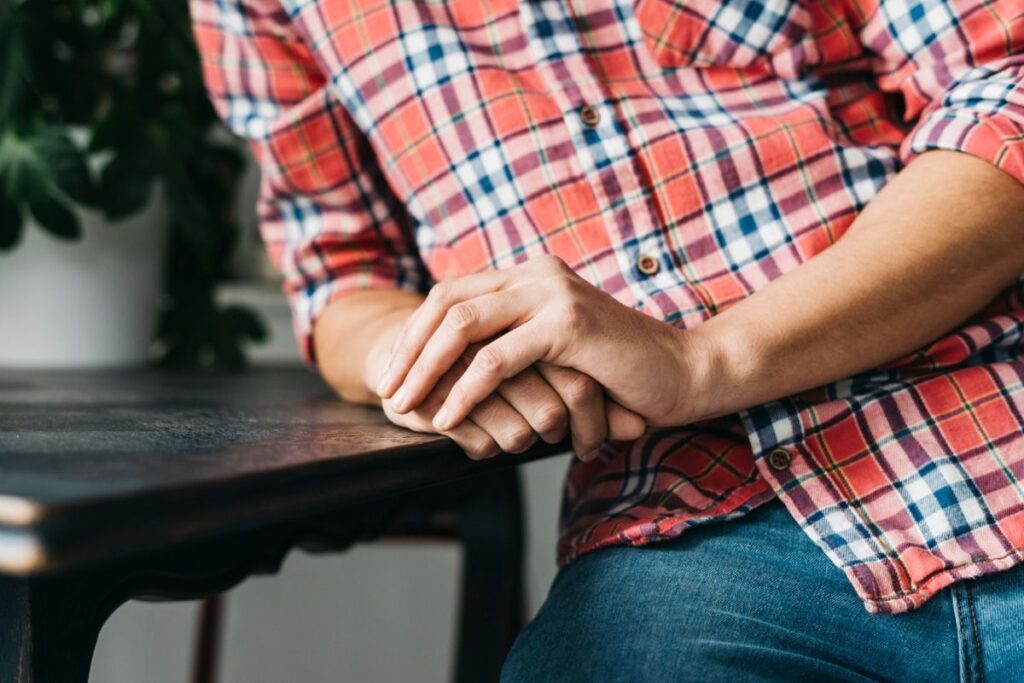 A person in a plaid shirt sits at a table, hands clasped together in front of them.