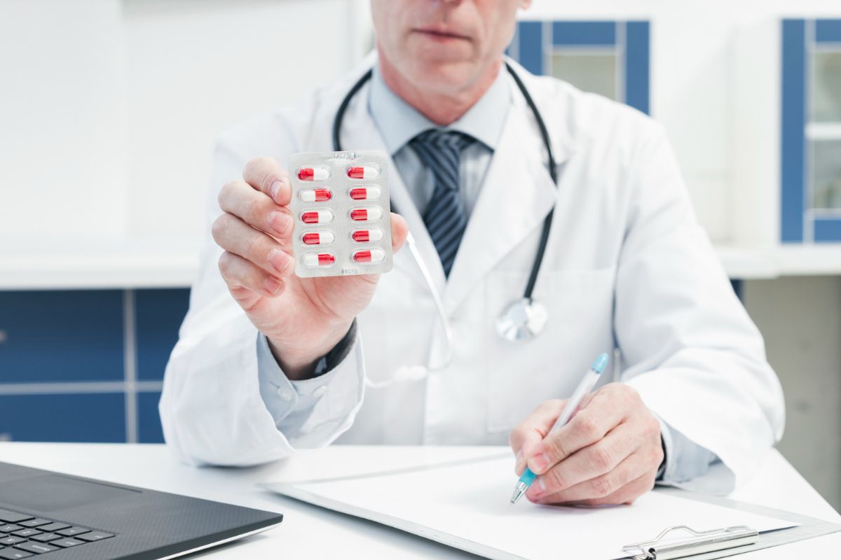 A male doctor in a lab coat holding a blister pack of pills, with a stethoscope around his neck and a laptop on his desk.