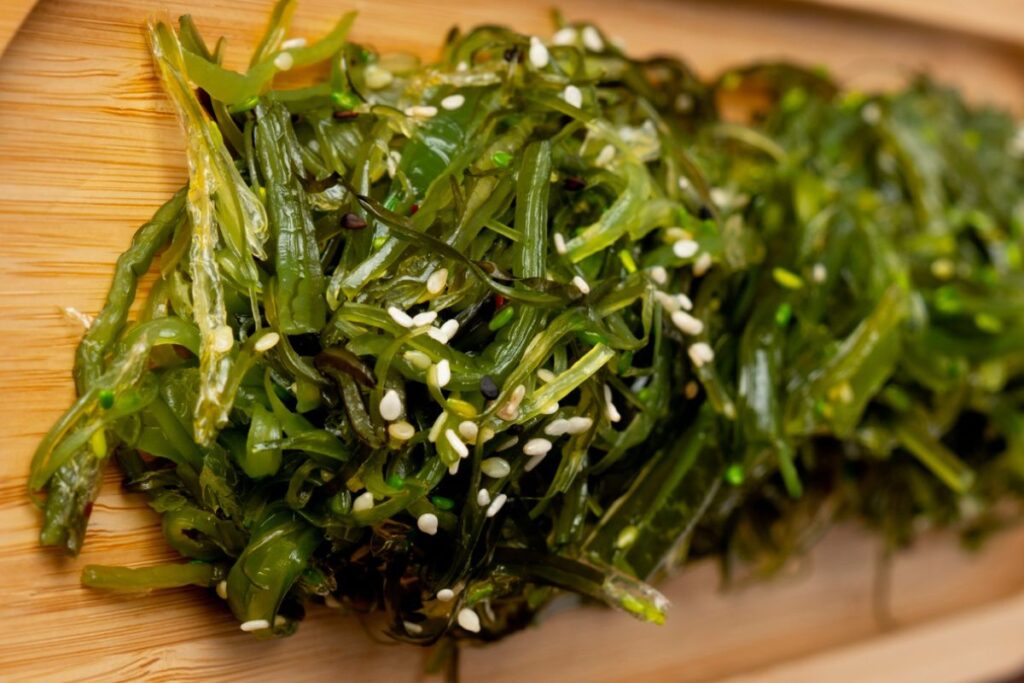 A close-up image of a serving of seasoned seaweed salad sprinkled with sesame seeds on a wooden plate.