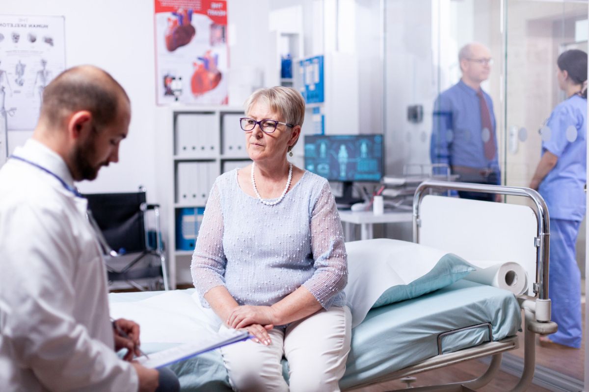 A senior woman sits talking to a male doctor in a hospital room, with a nurse and another healthcare worker visible in the background.