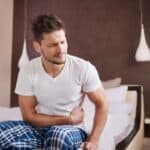 A man in pajamas sitting on the edge of a bed, clutching his stomach and grimacing in pain.