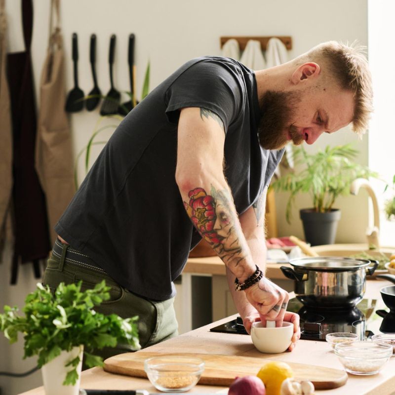 A bearded man with tattoos is cooking in a kitchen, chopping vegetables on a wooden board.He is busy preparing an anti-candida diet recipe.