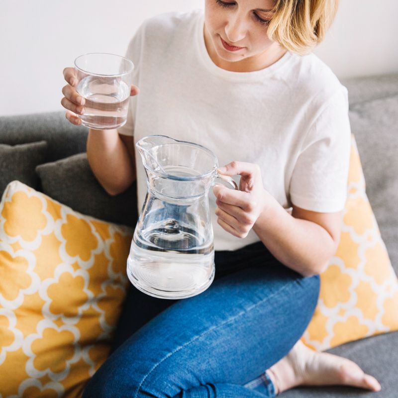 A woman sitting on a couch, pouring water from a pitcher into a glass.