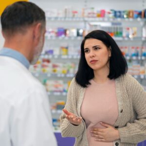 A woman, holding her stomach with a pained expression, speaks to a pharmacist in a pharmacy setting. Shelves with various medication boxes are visible in the background.