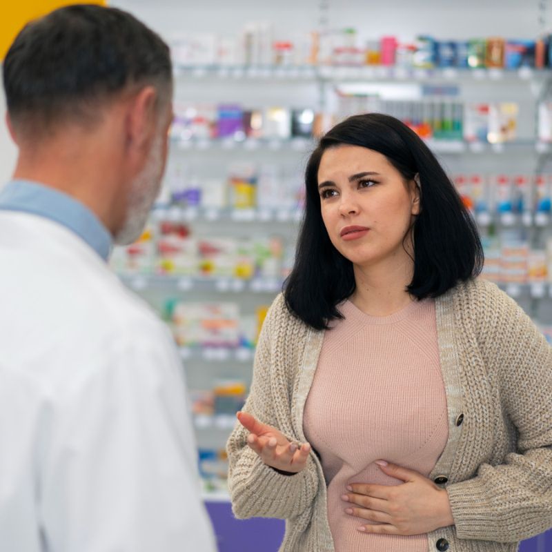 A woman, holding her stomach with a pained expression, speaks to a pharmacist in a pharmacy setting. Shelves with various medication boxes are visible in the background.