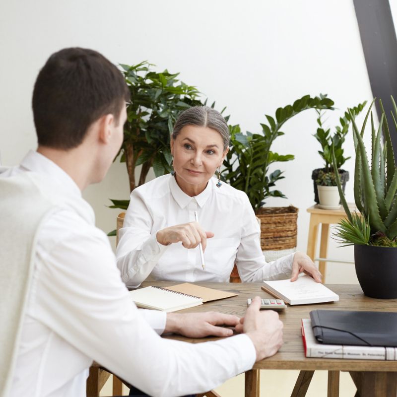 Two people sitting at a wooden table in an office setting, conversing. The background includes several plants, and both individuals have notebooks and pens in front of them.