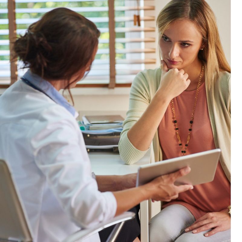 A woman consults with a doctor in an office. The doctor holds a tablet, and the woman listens attentively.