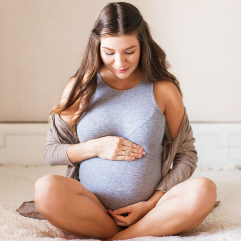 A seated pregnant woman with long hair smiles, looking down and holding her belly with both hands. She wears a gray tank top and a light cardigan, sitting on a bed with a light blanket.