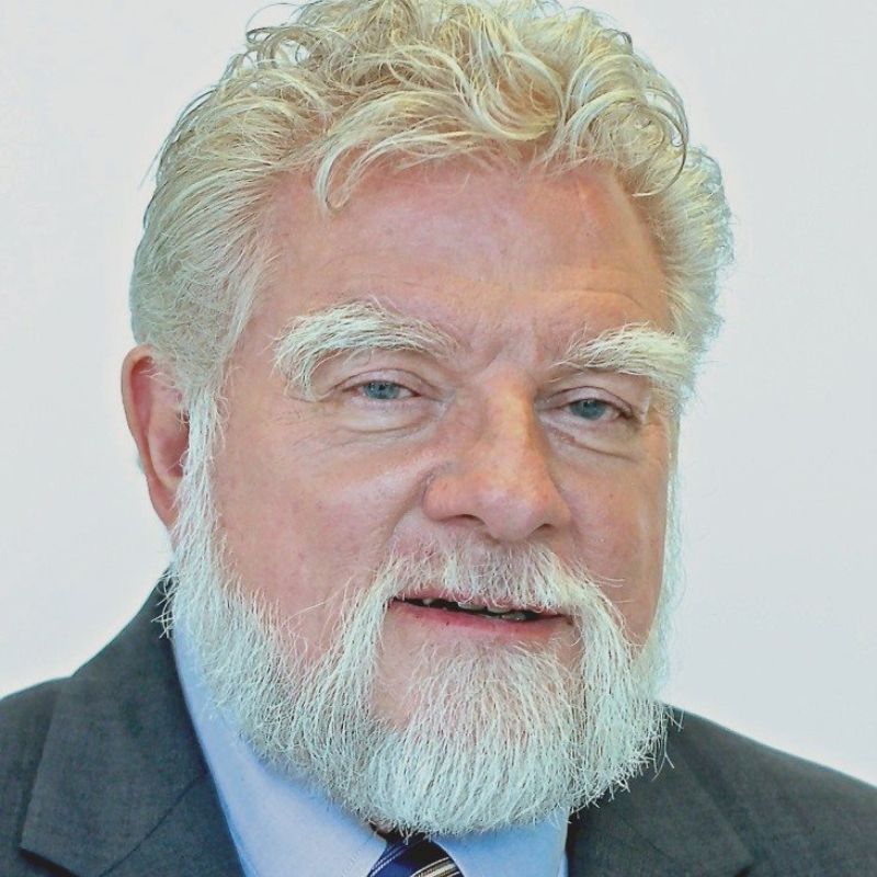 An older man with white hair and a full white beard, wearing a dark suit, light blue shirt, and striped tie, looking directly at the camera against a plain background.