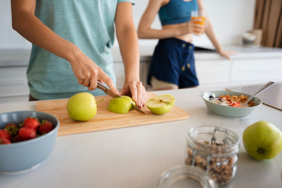 Two people in a kitchen, one slicing green apples on a wooden cutting board, and the other holding a glass of juice. A bowl of strawberries, a salad, and jars of granola are on the counter.