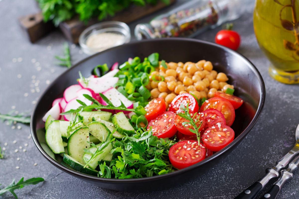 A colorful salad in a black bowl contains cherry tomatoes, chickpeas, cucumber slices, radish, sliced spring onions, and arugula, garnished with sesame seeds. A bottle of olive oil is nearby.