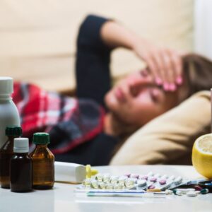 A woman lies on a couch with a hand on her forehead, surrounded by medication bottles, pills, and a lemon on a nearby table.