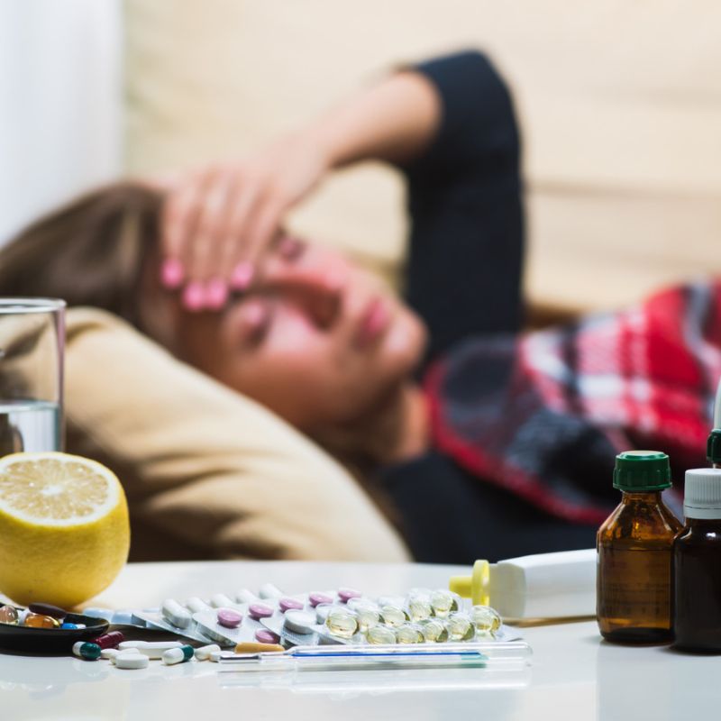 A person lies on a couch with a hand on their forehead, surrounded by various medicines, pill bottles, a glass of water, and a sliced lemon on a table in the foreground.