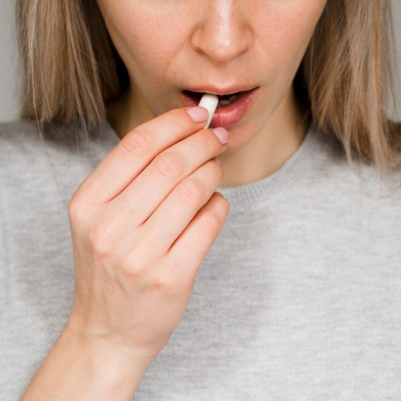 A person wearing a light gray top holds a pill near their mouth with one hand.