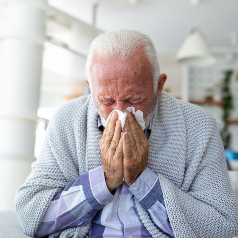 Elderly man with white hair and beard sneezes into a tissue. He's wearing a gray sweater and a checkered shirt, sitting indoors.