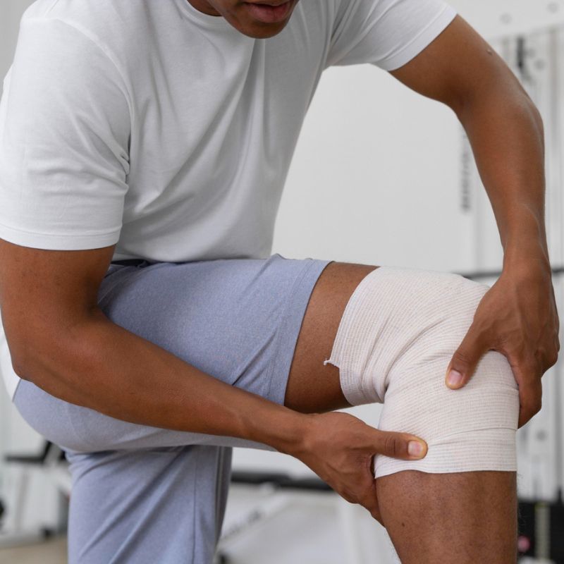 A person wearing a white t-shirt and gray shorts is adjusting a bandage wrapped around their knee.