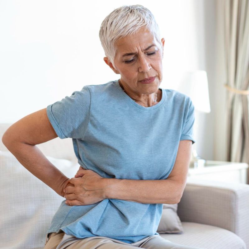 An elderly woman with short gray hair holds her back in pain while sitting on a couch, wearing a light blue t-shirt in a well-lit room.