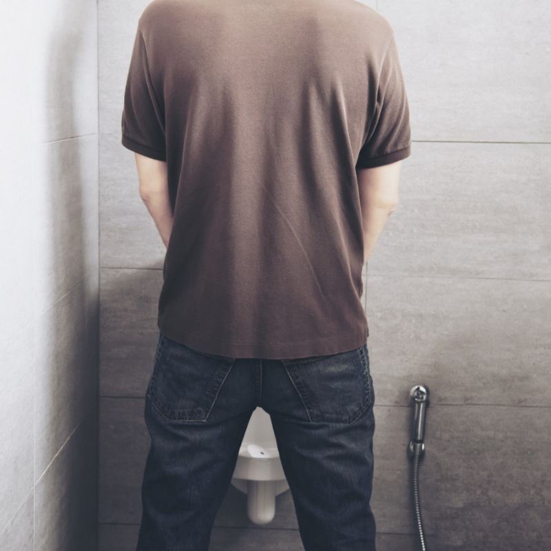 A person wearing a brown t-shirt and jeans stands facing a wall-mounted urinal in a gray-tiled restroom.