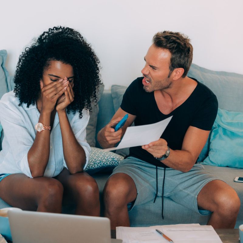 A man angrily points at a document while a woman covers her face with her hands, sitting together on a couch with a laptop and papers around them.
