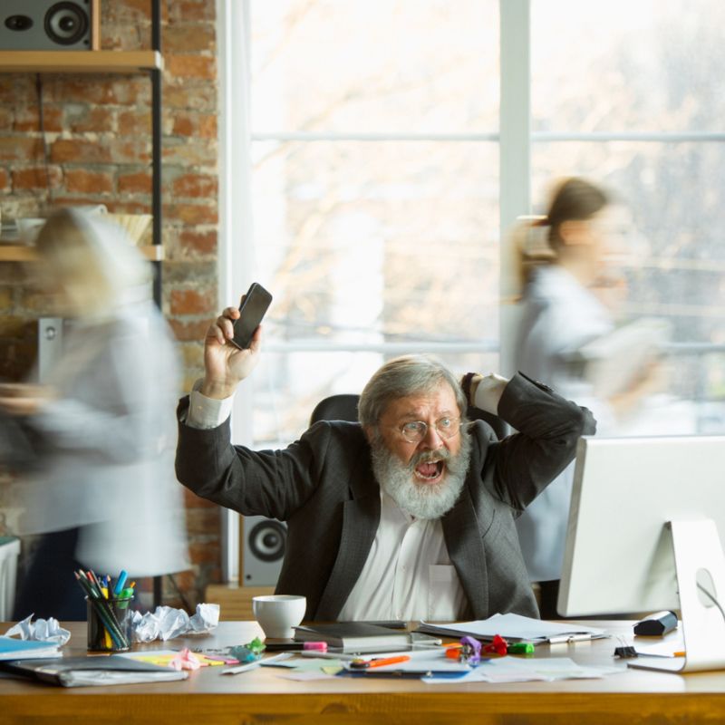 An older man with a beard appears frustrated, holding a phone and yelling at a cluttered desk, while blurred people move quickly in the background in a busy office setting.