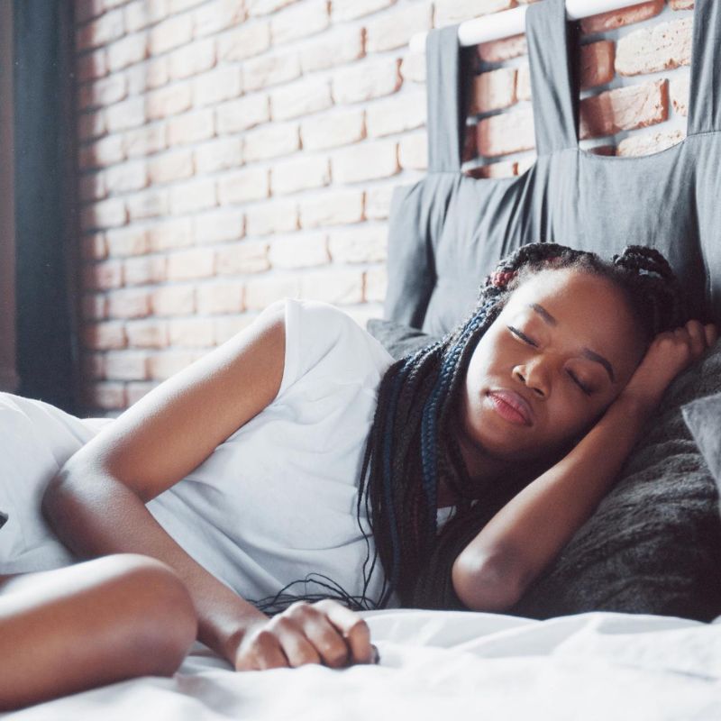 A woman with braided hair is lying on a bed, resting her head on a pillow, and appears to be sleeping. She is dressed in a white short-sleeved shirt, and the bed has white sheets and a brick wall backdrop.