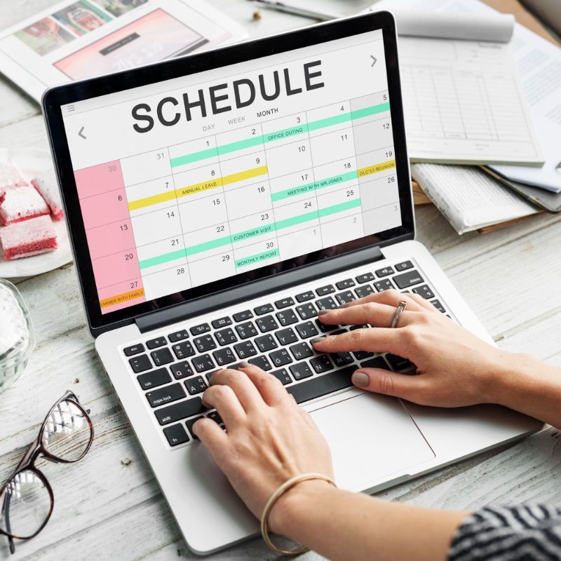Hands typing on a laptop displaying a calendar schedule. Nearby, there are a notebook, a book, and a cup of coffee on a white desk.