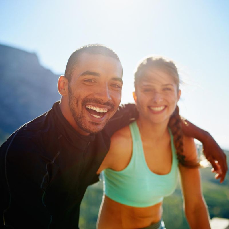 Two people, one in a black top and the other in a light green sports bra, smiling outdoors with a mountainous background and bright sunlight.