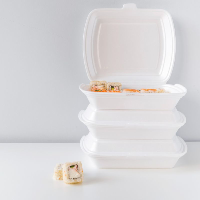 Three stacked white takeout containers with the top one open, revealing sushi inside. One piece of sushi is placed next to the containers on a white surface.