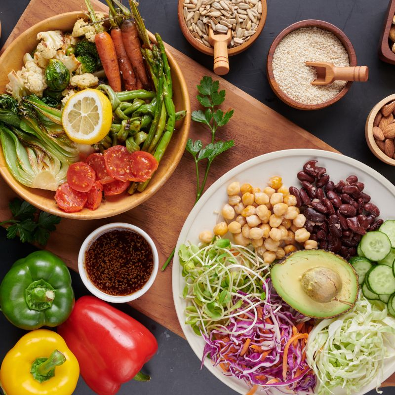 A tabletop filled with a variety of fresh vegetables, seeds, beans, and nuts. A wooden bowl contains carrots, asparagus, tomatoes, and more. A white plate has chickpeas, avocado, cabbage, and cucumbers.