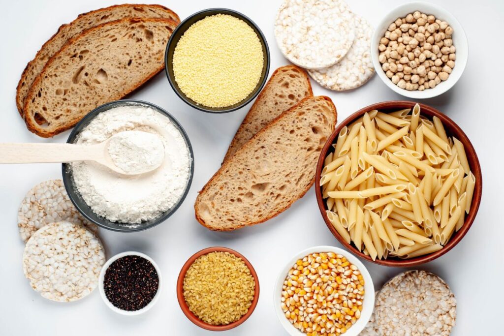 A variety of gluten-free foods including pasta, flour, bread, rice cakes, millet, quinoa, corn, and chickpeas are displayed in bowls and on a white surface.