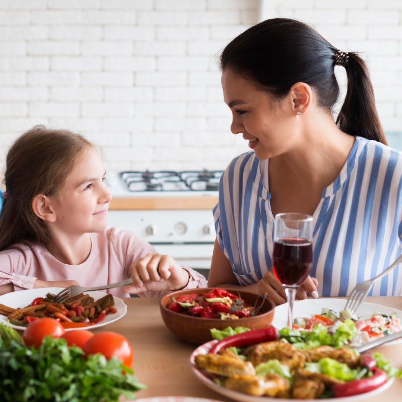 A woman and a girl smile at each other while sitting at a table with various dishes, including salad, vegetables, and a glass of red wine, in a kitchen setting.
