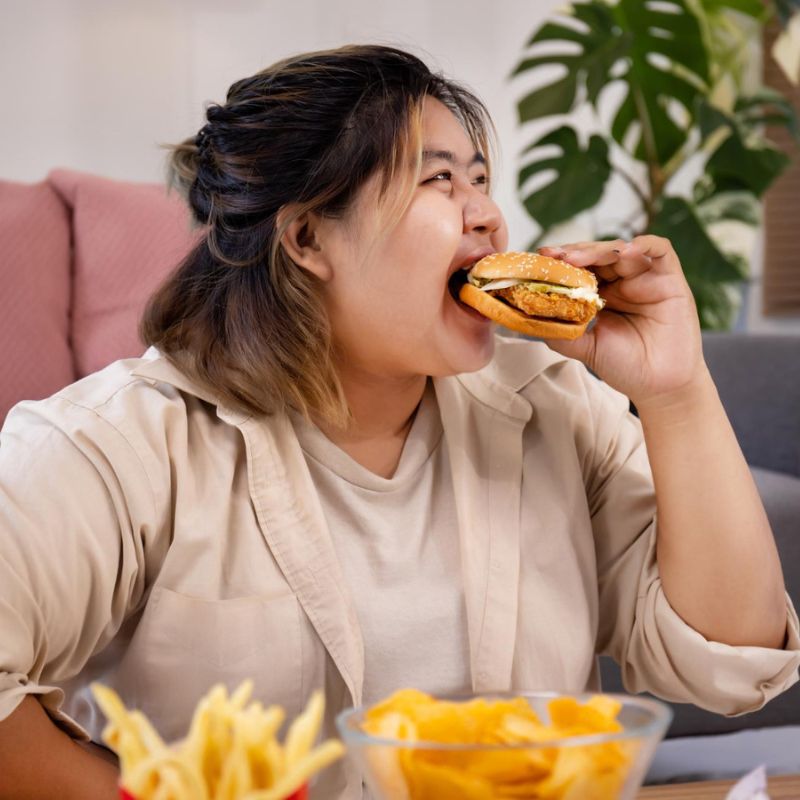 A person is sitting on a couch and eating a sandwich. There are bowls of fries and chips on the table in front of them.