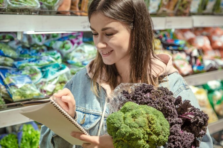 A person stands in a grocery store aisle holding broccoli and purple kale while looking at a candida diet food list and free shopping guide. Packaged greens are visible on the shelves behind them.
