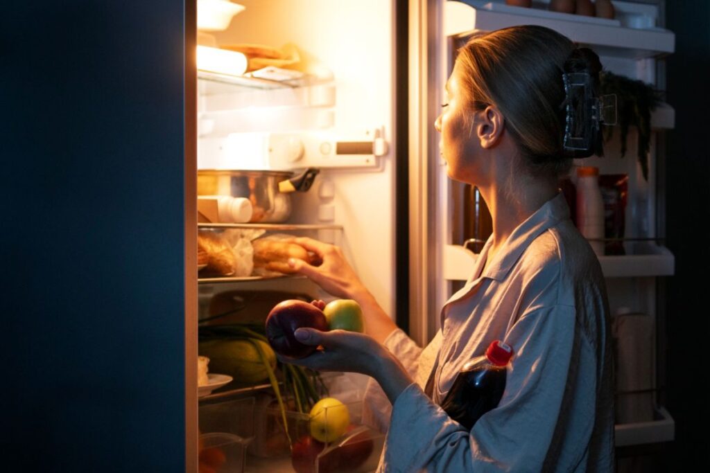 A woman in a robe looks into an open refrigerator, holding an apple, a peach, and a bottle of soda in her hands.