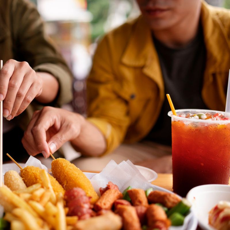 Two people in yellow jackets reaching for food on a table, including fries, chicken strips, and a large iced tea. Their faces are partially out of the frame.